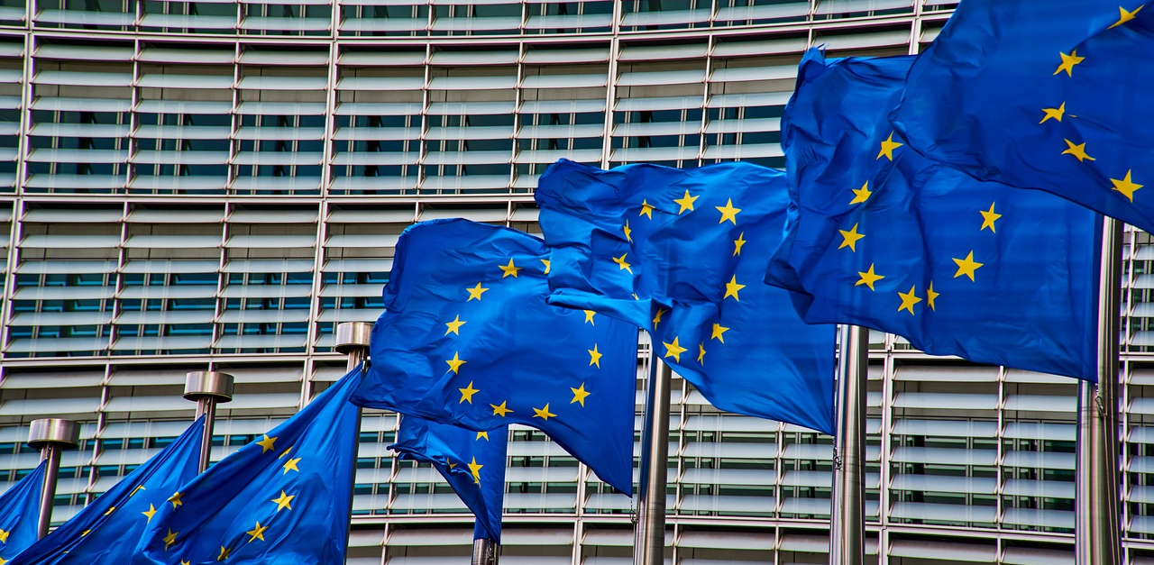 The image shows eight EU flags in front of the Berlaymont building in Brussels, which houses the headquarters of the EU commission.