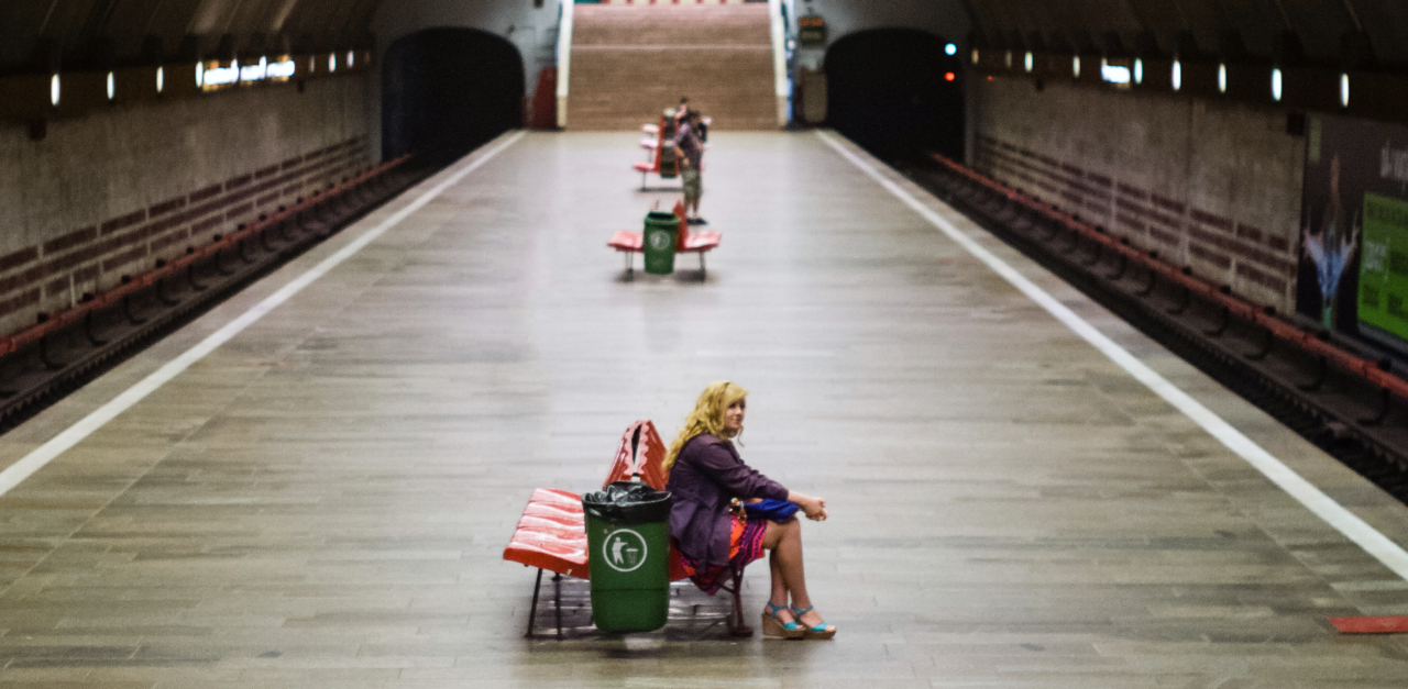 Photograph of the Titan Metro Station in Bucharest. It's taken from the side of a large tunnel, showing the long platform and some passangers waiting for trains. Train tracks run on boths sides of the platform. A woman sitting in the foreground of the photo is in focus and the background is slightly blurred.