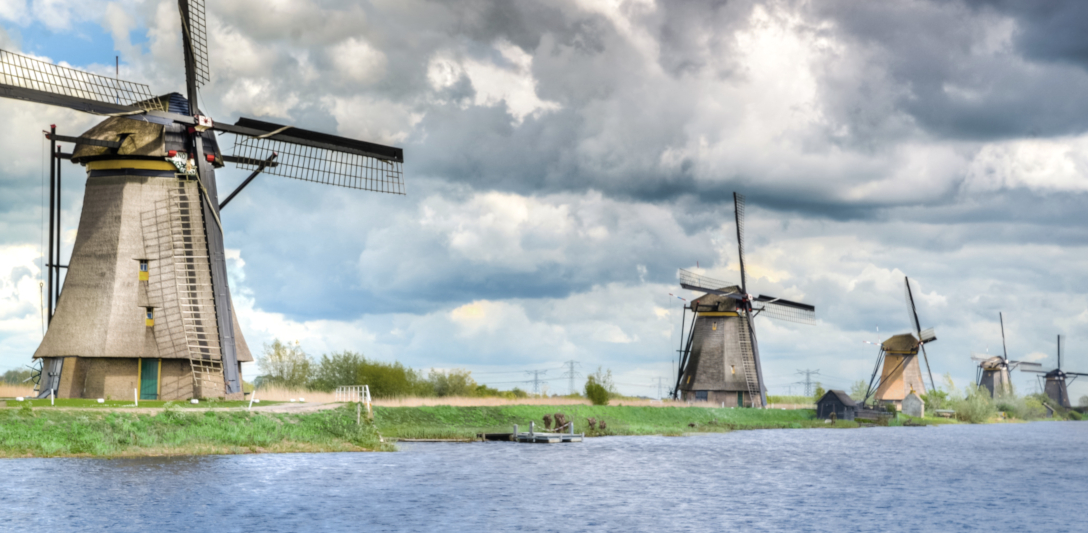 Photograph of five windmills on a grassy field next to water. The photo is taken from a perspective where the first mill looks significantly bigger than the second one, etc, until the last windmill in the distance. There sky above is filled with clouds.