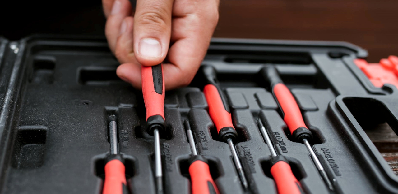 Photograph of a hand grabbing a screwdriver from out of a toolbox or toolset that is open on a table. There are several similar screwdrivers lying next to the one being held by the hand.