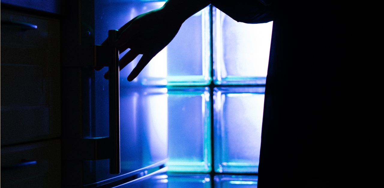 The siluette of a torso and a hand reaching for a refrigerator handle, in black against a backdrop of blue light shining through a glass wall.